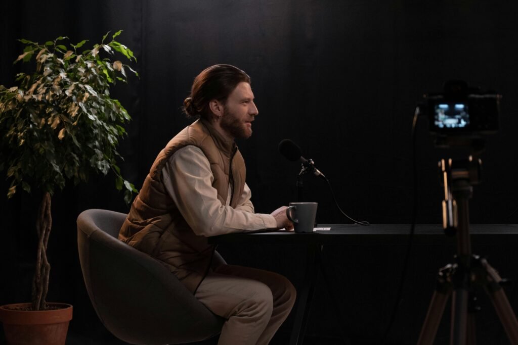 A man sits at a table in a studio setting, recording a podcast with a microphone.